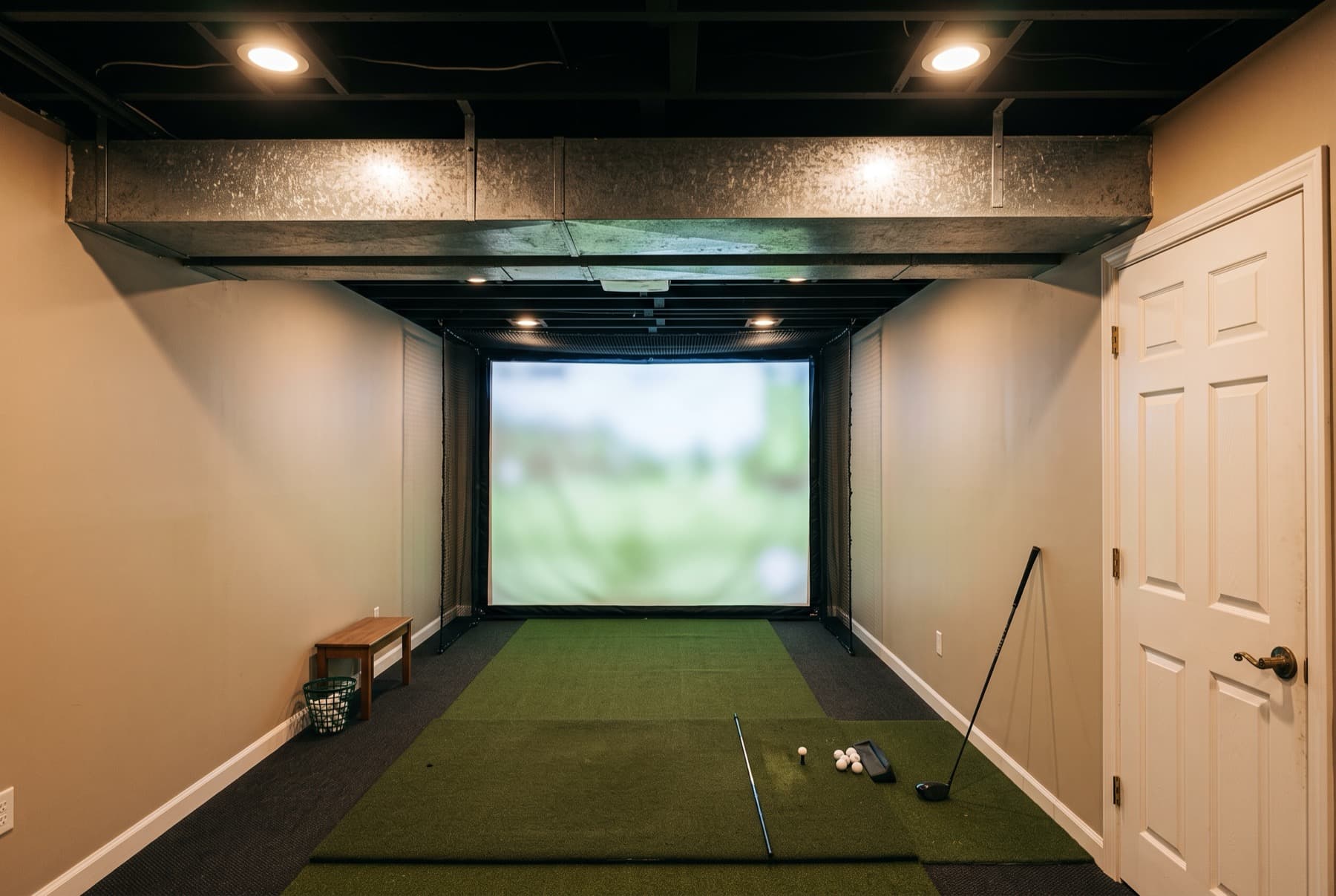 A basement golf room where a galvanised duct hangs below the joists, directly over the hitting mat.